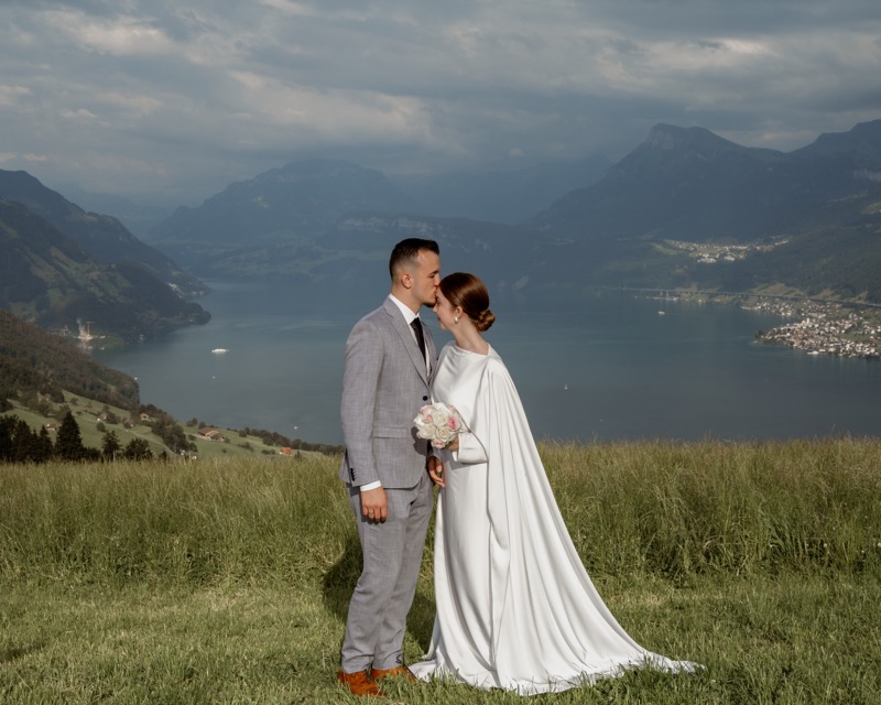 Bräutigam küsst die Braut auf die Stirn, dahinter See und Berge
