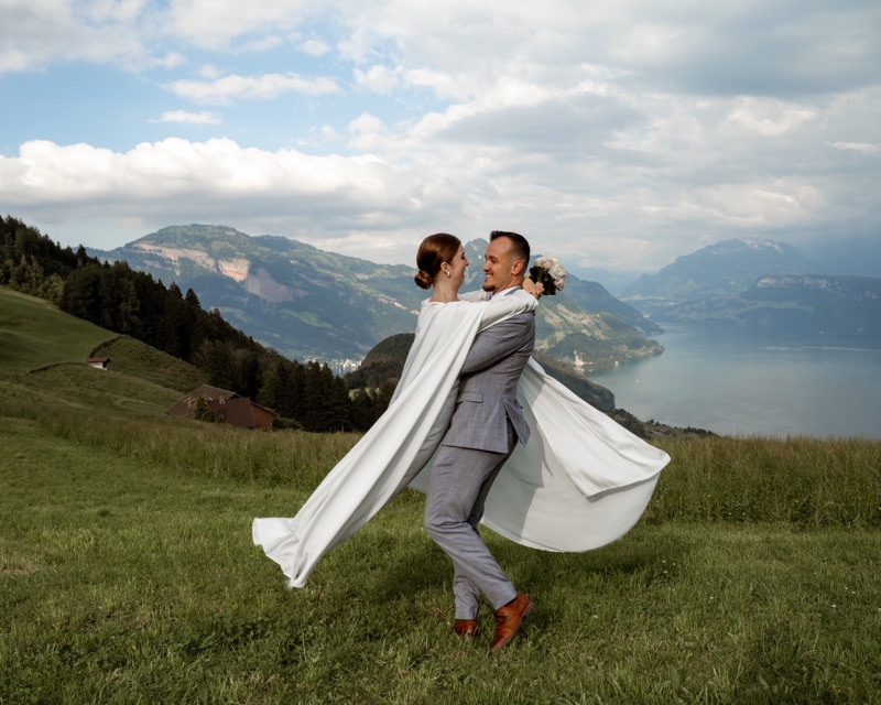 Bräutigam hebt die Braut hoch, ihr Umhang weht über der Bergwiese